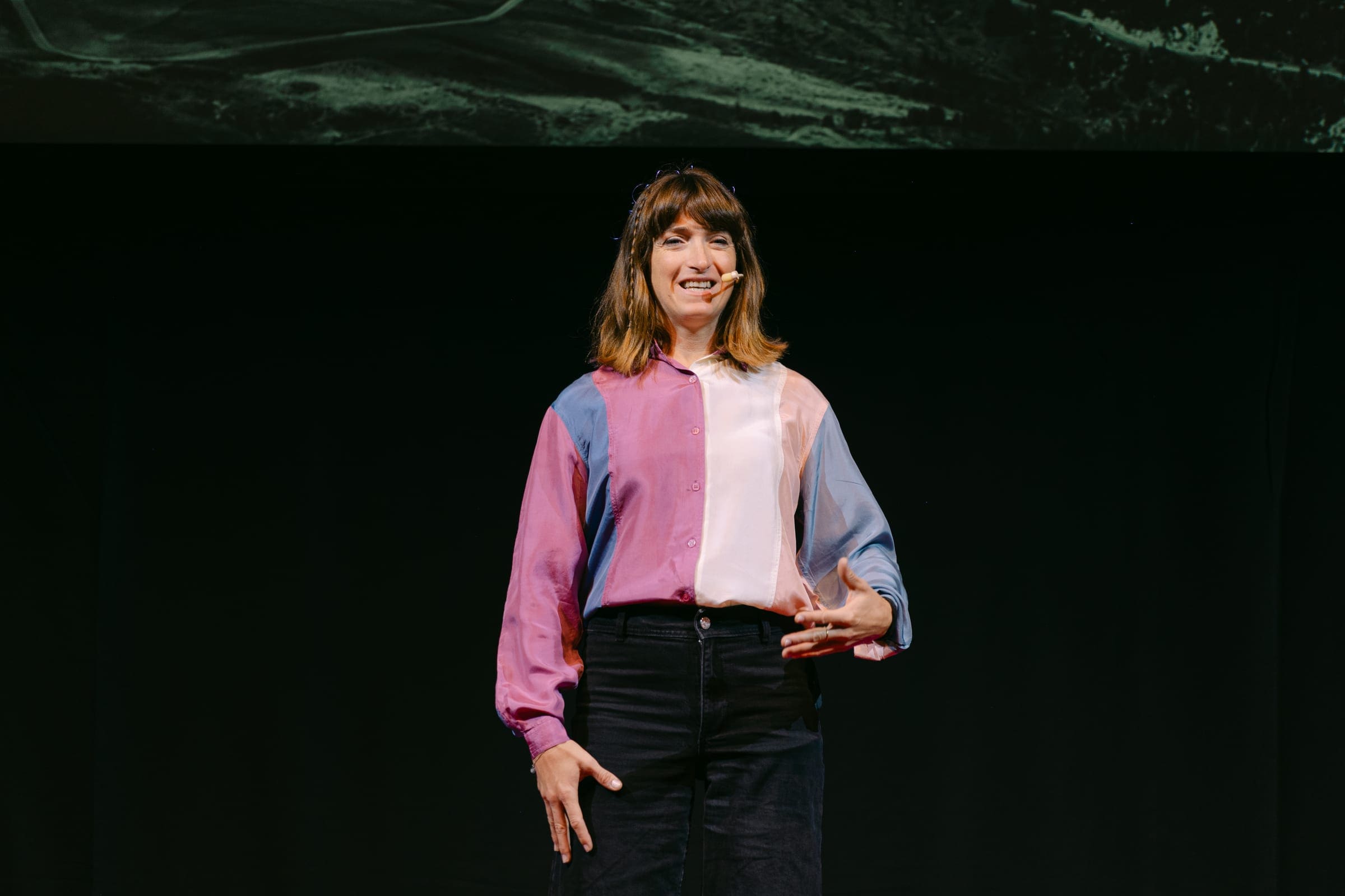 Martina Caironi speaks on the TEDxCortina 2025 stage wearing a headset microphone and a colour-block shirt